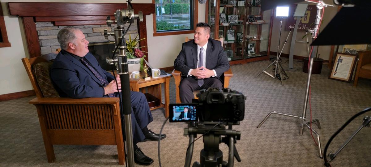 Two men speaking to each other for a podcast in a well-lit room surrounded by lights, a camera, and other audiovisual recording equipment.