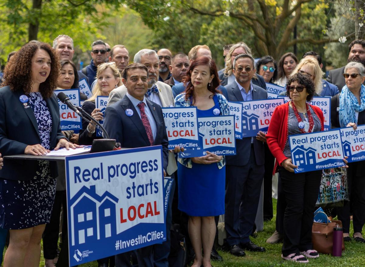 A diverse group of city officials holding signs that say "real progress starts local" looks at a speaker.