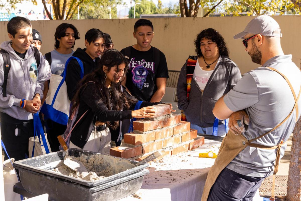 A high school student learns how to lay bricks, as a other students and a teacher.
