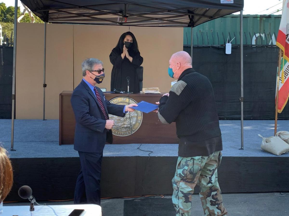 A man handing another man a folder in an outdoor courtroom with a judge looking on.