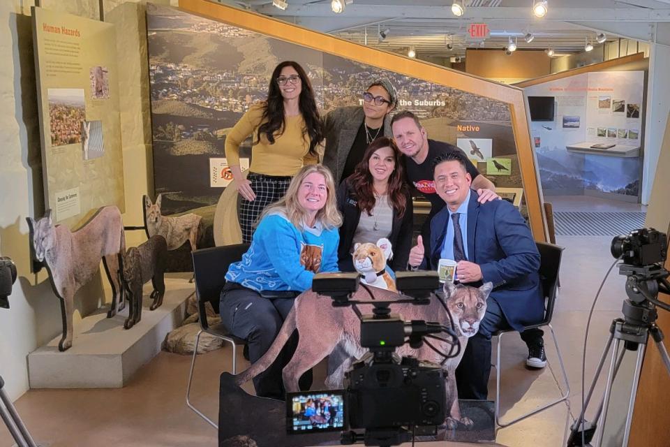 A group of men and women posing for a picture next to podcasting equipment and cardboard cutouts of wildlife.